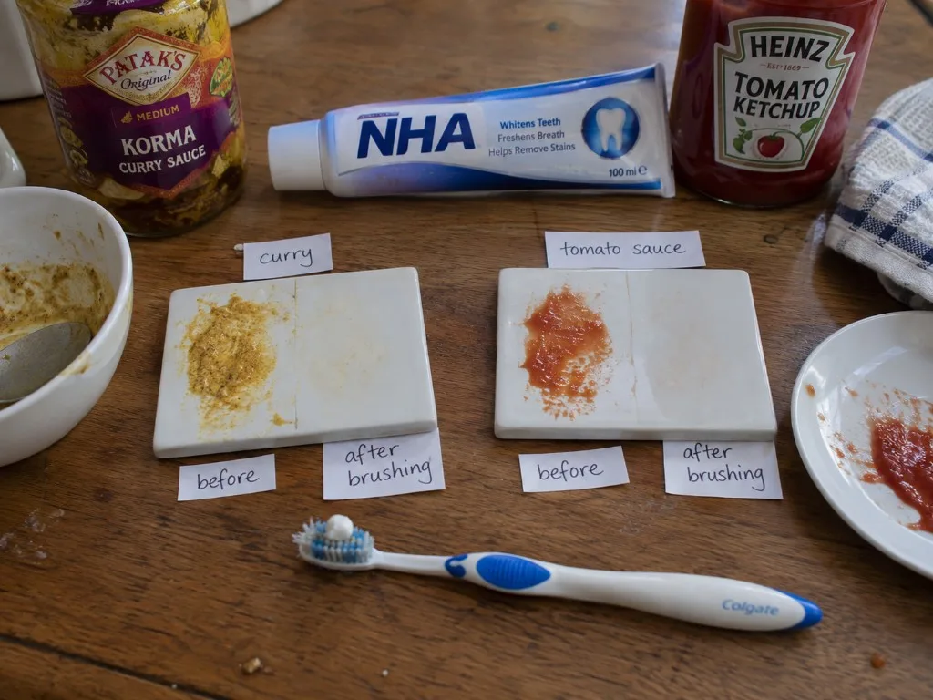 Nano-hydroxyapatite toothpaste stain wipe: curry tile split before and after brushing, red sauce tile split before and after brushing, with the nHA tube, a Colgate brush, Patak's korma, and Heinz ketchup